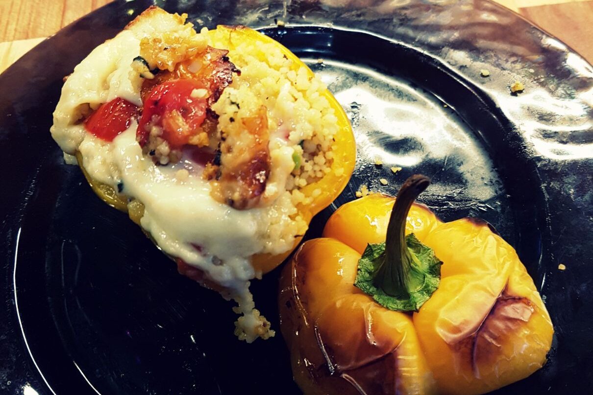 A plated stuffed yellow bell pepper topped with melted cheese and couscous, served on a purple glass plate.
