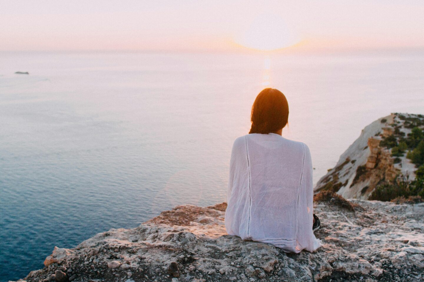 Woman sitting peacefully on a cliff at sunrise, meditating in nature and symbolising gentle ways to nurture mental health and wellbeing.